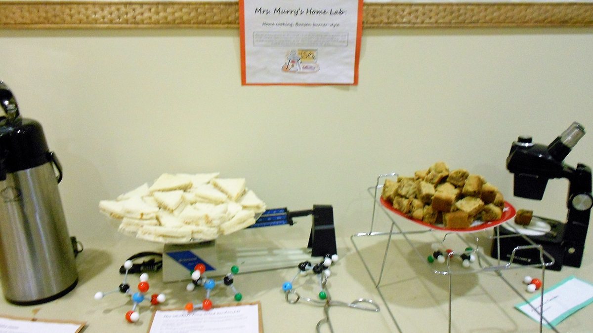 Buffet table with hot drink dispenser, tuna sandwiches on a scale, cookies on a wire rack, and a stereo microscope, labelled "Mrs. Murry's Home Lab"