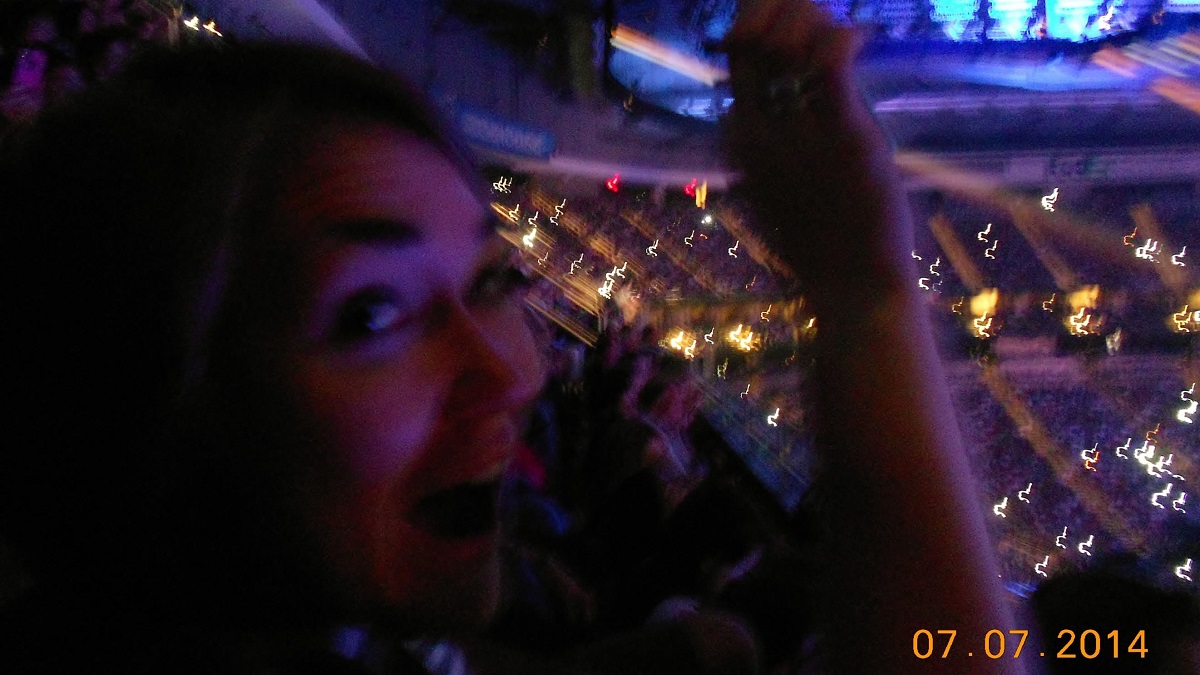 Excited woman waving a lit cell phone (the modern-day lighter) amid an arena full of other people, other lit cell phones speckling the crowd.