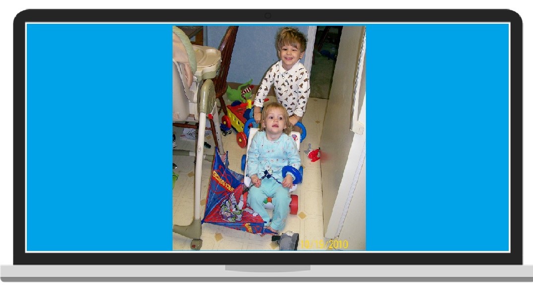Small child pushing his 18mo sister, who is carrying an open umbrella and wearing stacking rings, around a messy kitchen in a push toy like a stroller.