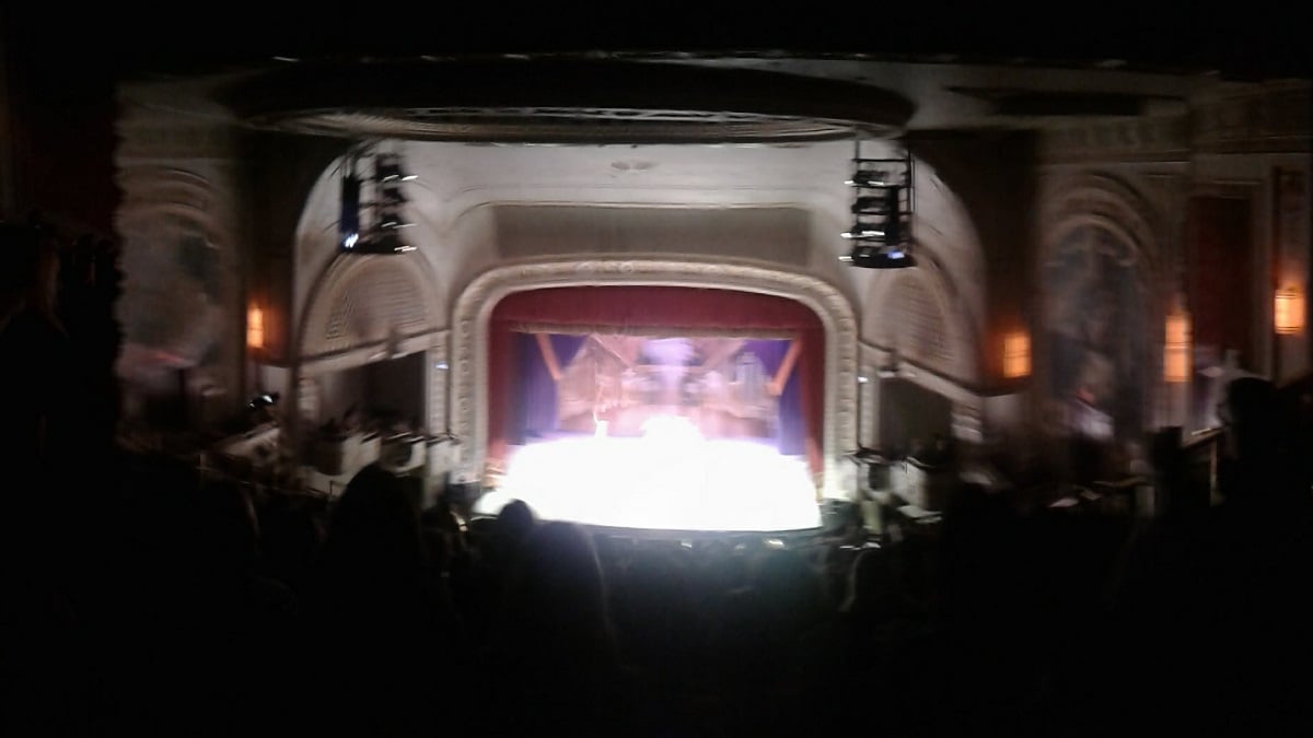 Panorama view from the highest rows of a theater, the stage in the center so brightly lit as to wash everything on it out