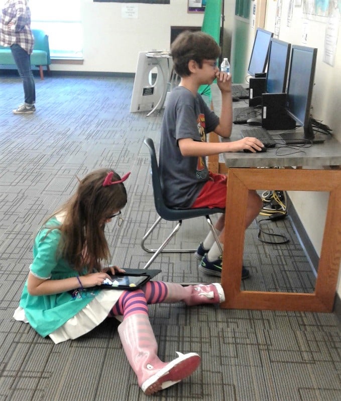Girl in Star Butterfly costume sits on floor with a tablet beside boy at a table playing on a computer