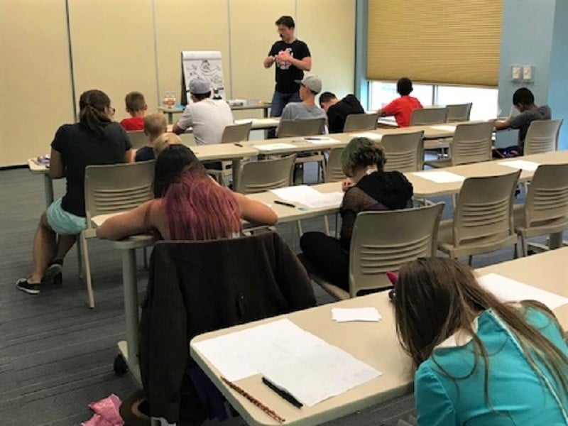 Man stands before tables of a dozen kids busy drawing